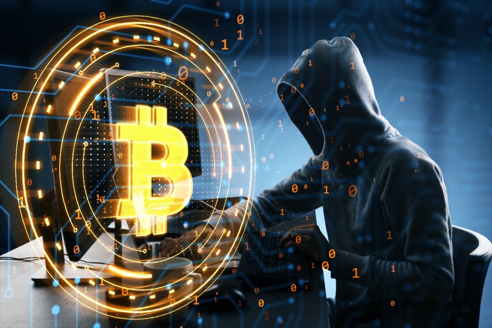 Fraudes digitais e medidas do BC podem acelerar a chegada de regras para VASPs. Hacker de capuz manipulando um computador com um grande símbolo de Bitcoin brilhando em primeiro plano