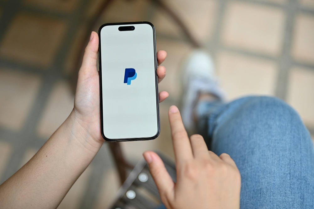 Pessoa segura um smartphone exibindo o logotipo da PayPal, representando o uso de criptomoedas em pagamentos online.