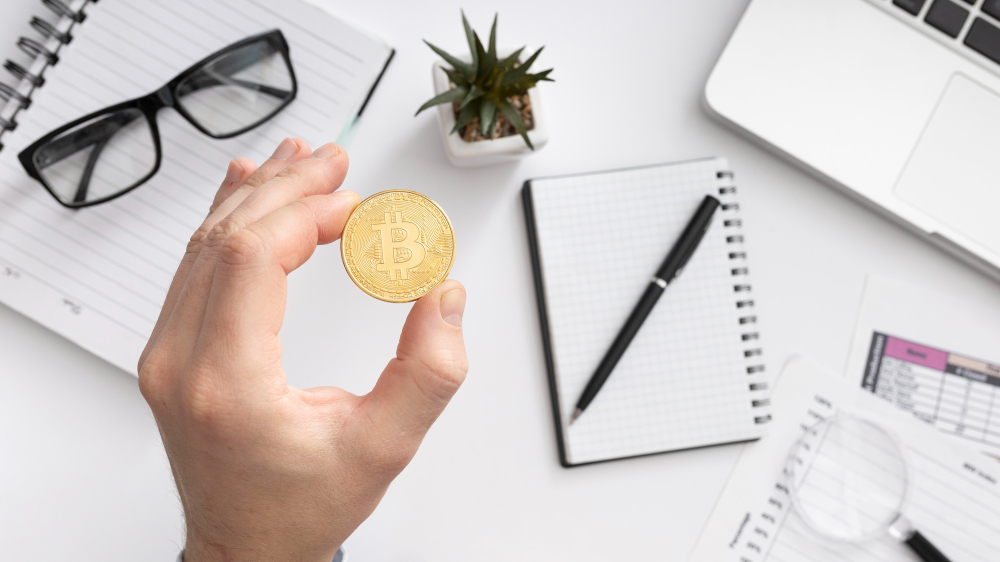 mao segurando moeda de bitcoin sobre mesa de estudos simbolizando educacao financeira e introducao das criptomoedas no ambiente escolar